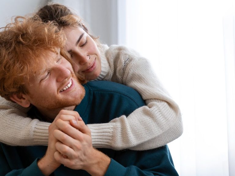 Un couple souriant, l'homme assis, la femme l'enlace tendrement.